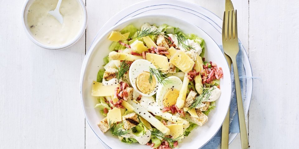 Een rijkgevulde salade staat klaar op tafel. Het witte bord is gevuld met sla, gekookte eitjes, stukjes asperges en kaasschilfers. Dat is de lente proeven in je bord, als je het mij vraagt!