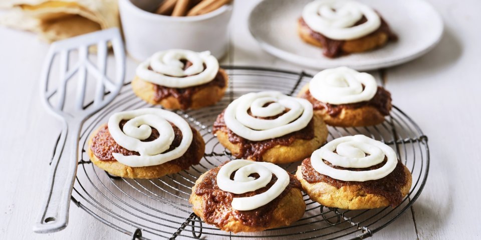 Op een taartrooster liggen 6 koekjes af te koelen. Het zijn koekjes van cinnamon rolls, een nieuwe versie om te genieten van deze heerlijke en kruidige zoetigheid. Op de koekjes is frosting gespoten op basis van roomkaas. 