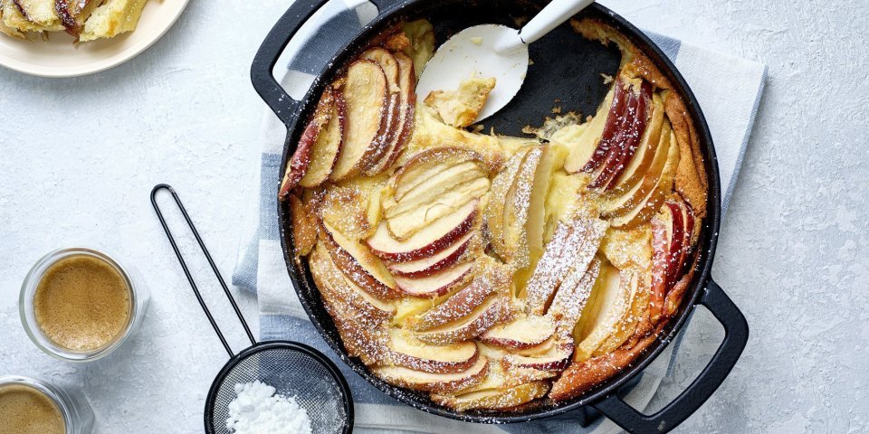 Een clafoutis van appelstukkjes staat op het aanrecht in een zwarte fonte schaal. Er is al een portie uitgeschept en staat op een bordje naar de pot. Er is ook bloemsuiker over het dessert gestrooid en de koffie is al uitgeschonken. 