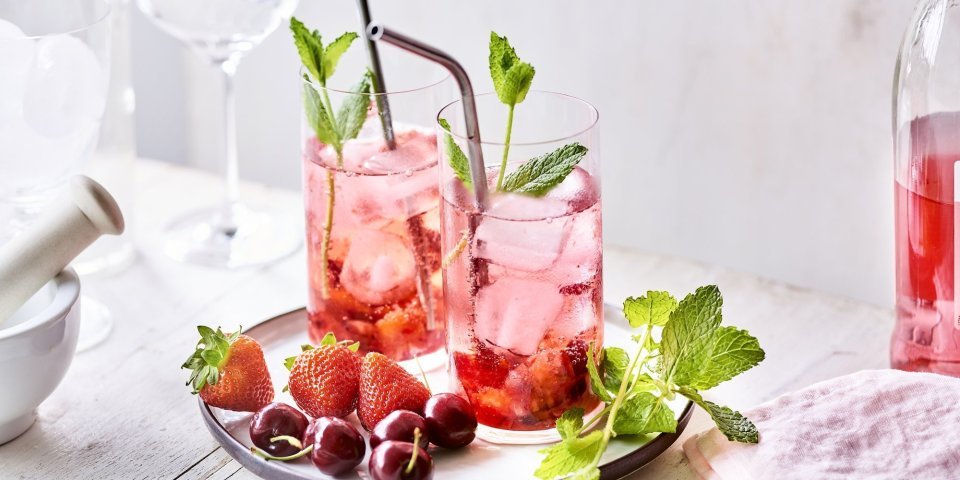 Twee cocktails staan op tafel, ze hebben een mooie roodroze kleur. Onderaan het glas ligt het smached fruit, met daarna de vodka en roze tonic. Er liggen nog enkele aardbeien en kersen langs. 