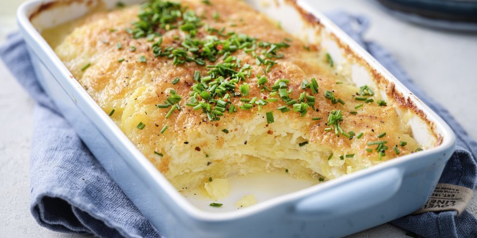 De ovenschotel met gratin staat op tafel, hij heeft een lichtblauwe kleur. De aardappelschijfjes zijn lekker crispy gebakken, doordat we ze in de oven klaarmaken met minder room. De ovenschotel is afgewerkt met verse fijngehakte bieslook.