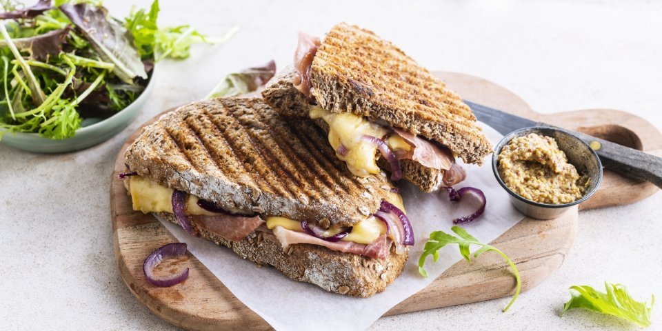 De croque ligt op een houten plankje aangesneden. De kaas en rode ui zien er heerlijk uit. In de achtergrond zie je ook nog wat gemengde sla en een potje graantjesmosterd. 