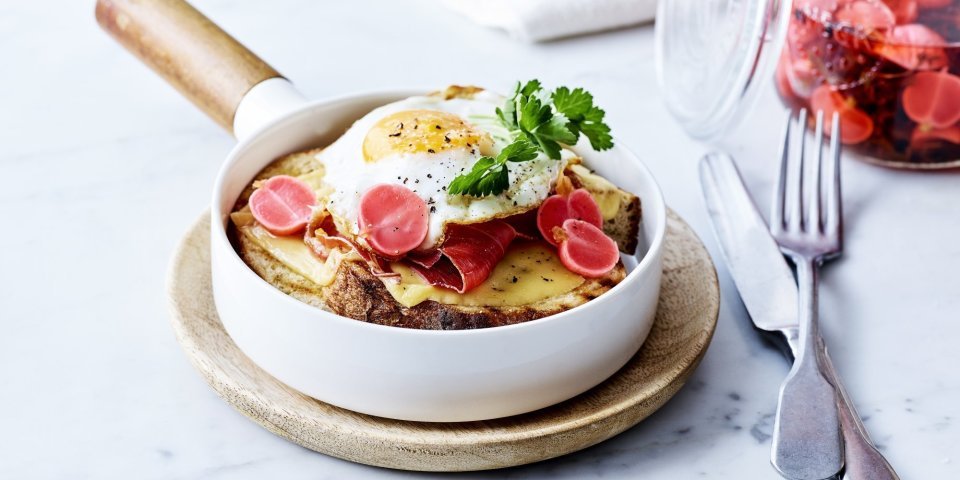 Croute au fromage met Averbode kaas in een witte pan, spiegeleitje er bovenop gepresenteerd met een takje verse peterselie. Op de achtergrond staan gepekelde radijzen in glazen pot.