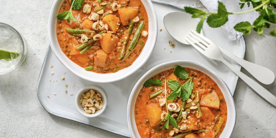 Twee heerlijke curry staan uitgeschept op tafel. Ze hebben een mooie oranje kleur door, jawel, de curry pasta. Ze staan beiden op een poseleinen dienplateau met bestek en een potje pina- en hazelnoten. 