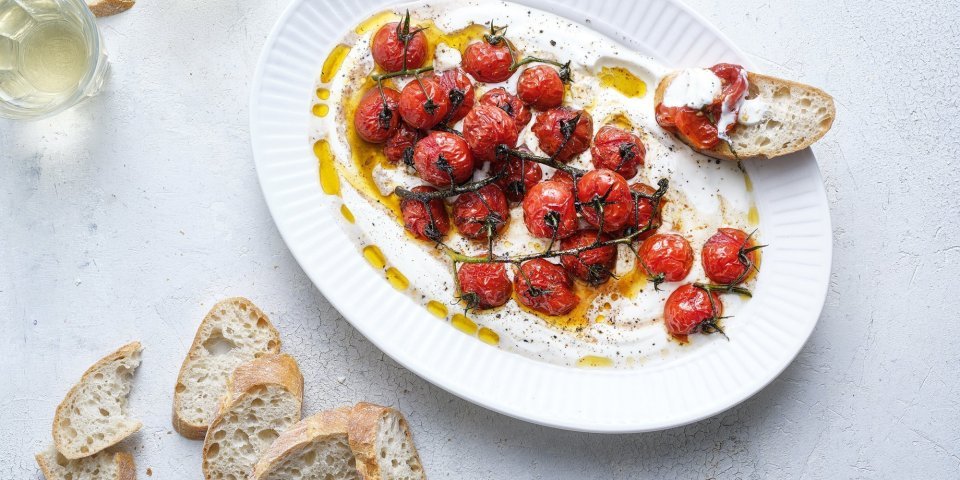 Een groot ovaal bord is gevuld met de romige yoghurt waarop de dieprode kerstomaatjes bovenop liggen. Er liggen enkele sneetjes stokbrood naast om te dippen. Er staan ook twee glaasjes witte wijn in beeld. 