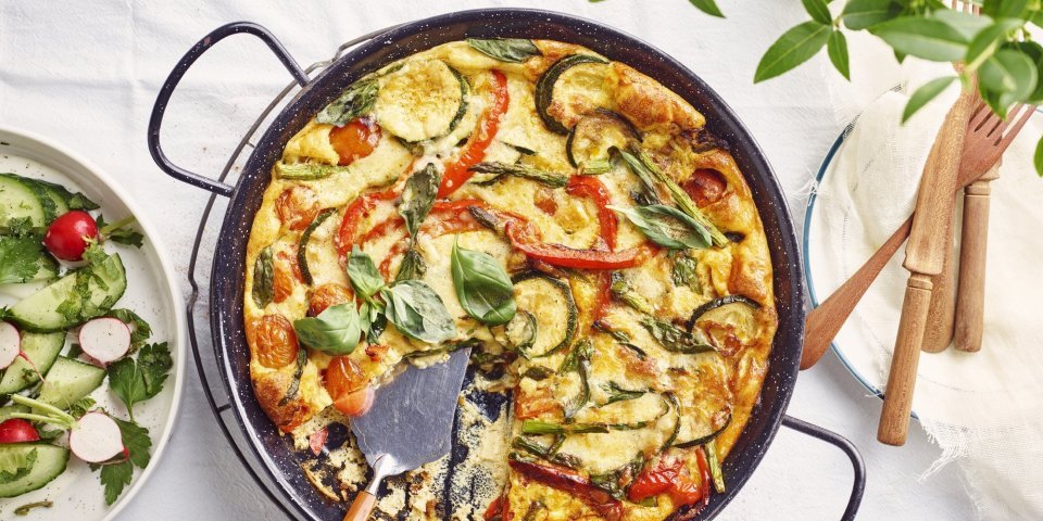 De frittata staat op tafel in zwarte pan, waardoor hij nog mooier oogt dan dat hij al is. De tomaatjes, paprika, courgette en asperges geven leuke kleurtjes aan de frittata. Er staat een slaatje langs de linkse zijde, met komkommer, radijsjes en verse kruiden.  