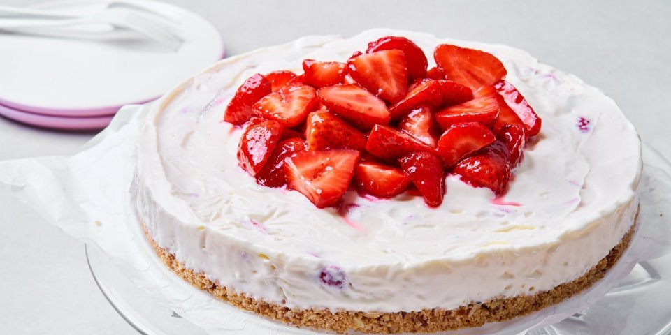 Een heerlijke cheesecake staat op een glazen serveerschaal. Dit is niet zomaar een cheesecake, maar eentje die diepgevroren is. Perfect voor een koel dessertje op een zomerse dag. Er liggen enkele verse aardbeien op de taart om het fruit extra in de verf te zetten, want aardbeien zijn gewoonweg de toppers van het zomerfruit. 