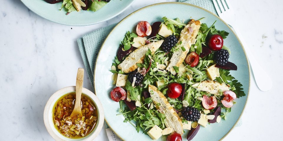 Een gemengde salade met sla, kersen, bramen en gekookte kipfilet gedresseerd op een lichtblauw bord. De dressing is er naast gezet in een keramieke potje met houten lepeltje in, de dressing bestaat uit sjalot, rode wijazijn, olijfolie en peper en zout. 