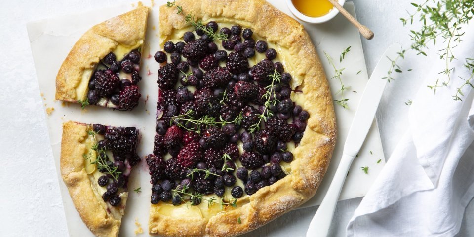 Galette met bramen, blauwe bessen en Belgische brie ligt aanhesneden op het aanrecht. Vergezeld met een potje gouden honing en een tijmplantje. 