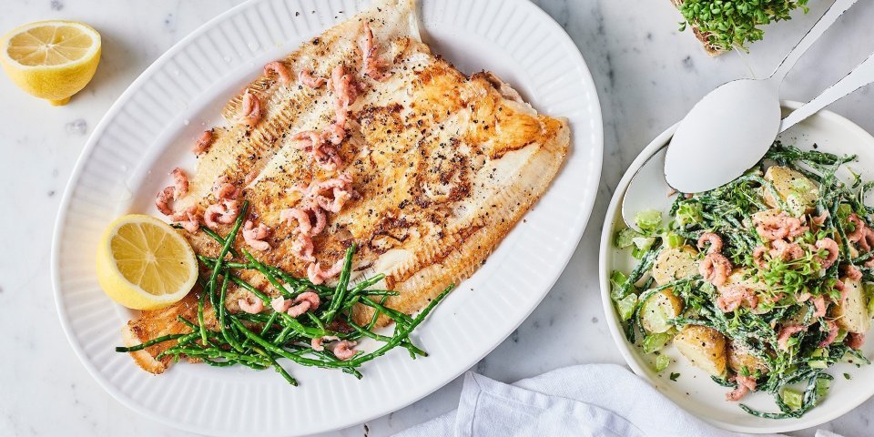 Een heerlijk krokant visje afgewerkt met zeekraal ligt op een groot bord. Er zijn enkele garnaaltjes aan toegevoegd die mooi roze blinken. De aardappelsalade staat er langs in een witte kom met witte lepel. Om af te werken ligt er 1 citroen doormidden gesneden.