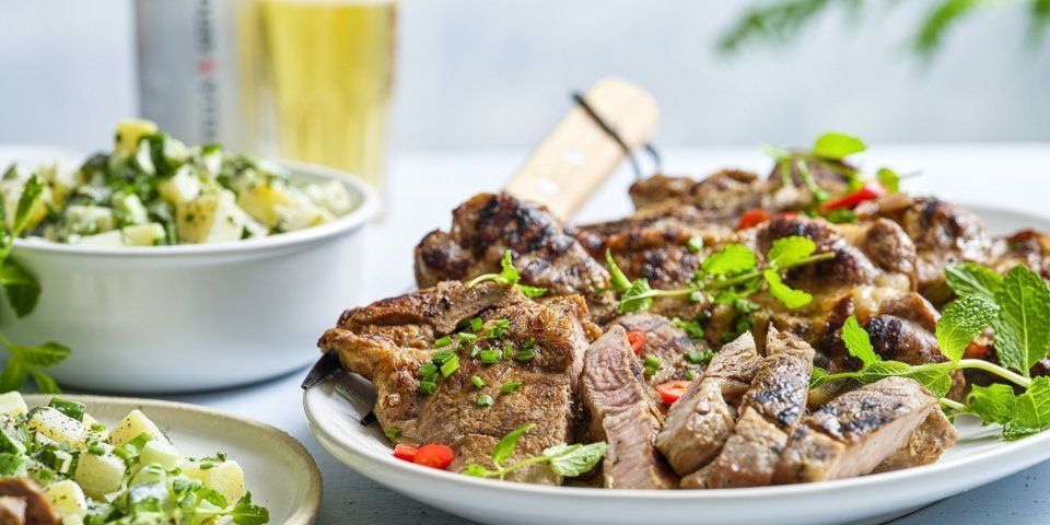 Een spieringkotelet ligt op een grote witte schaal in reepjes gesneden. Ze is mooi gegrild en afgewerkt met verse kruiden zoals munt, bieslook en chilipeper die voor extra kleur zorgen. Er is een aardappelsalade bij geserveerd alsook een frisse pint. 