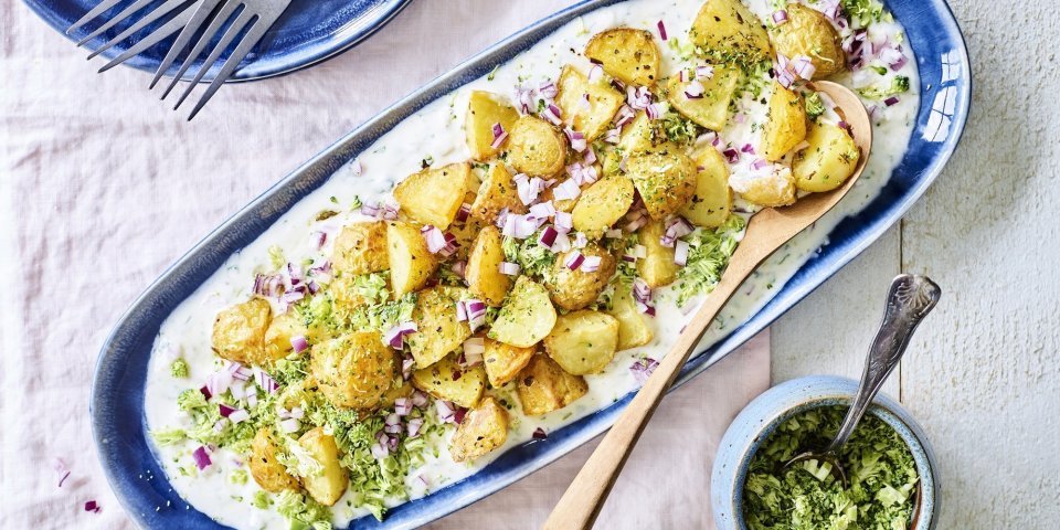 Een langwerpig blauw bord staat op tafel. Hierop is yoghurt op geschept met krokante aardappelen, stukjes rode ui en geraspte broccoli. 