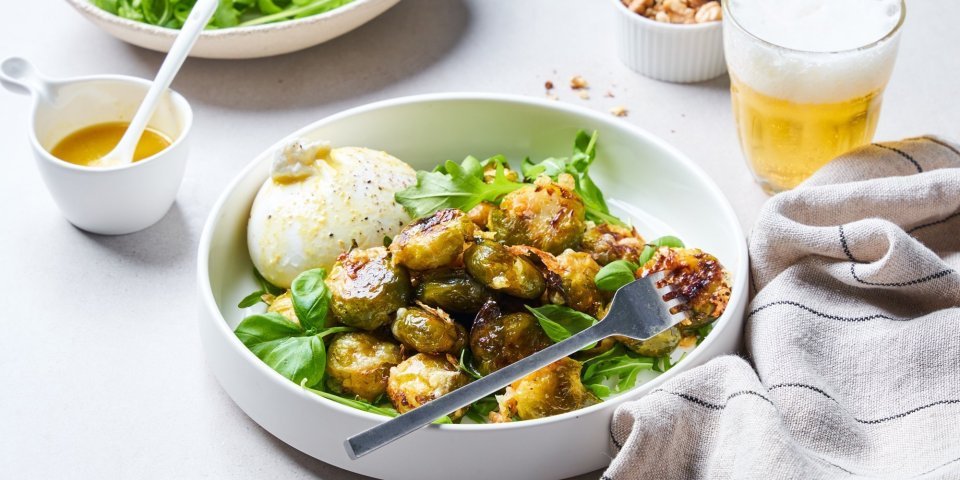 In een diep wit bord liggen de geroosterde spruitjes op een bedje van rucola en basilicum. Hierlangs ligt de burrata. Er is al wat vinaigrette gesprenkeld op de groenten en burrata.