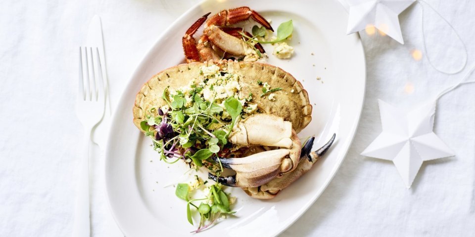 Als het feest is, dan mag het wat meer zijn toch? Gevulde krab met homemade krabsla ligt op een ovaal wit bord met wit bestek lang. Glaasje witte wijn staat bovenaan het bord en er liggen kerstlichtjes en -sterren mee op tafel.