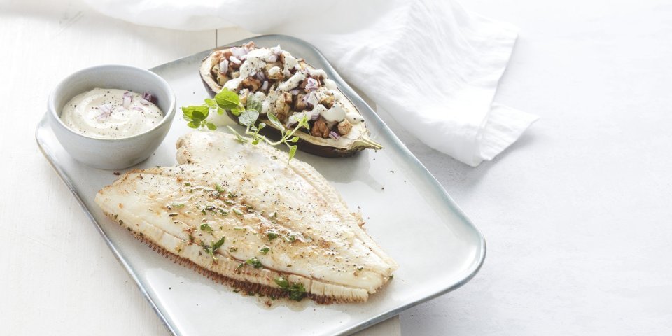 Een gebakken schartong ligt op een rechthoekig wit bord. De halve aubergine ligt naast de gebakken vis, hij is gevuld met stukjes knolselder, peer en walnoten. Het gerechtje is afgewerkt met verse gehakte kruiden en een lepeltje zure room.