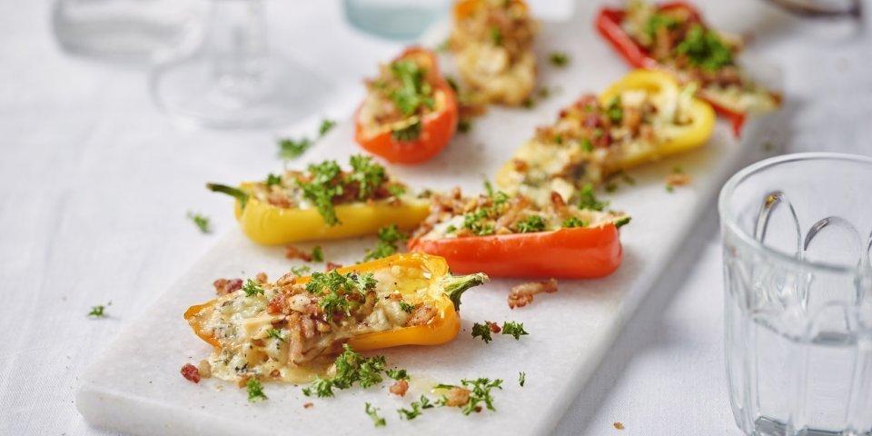 Gele en oranje snackpaprika's liggen op een witte stenen snijplank. De paprika's zijn gevuld met kaas, noten en crumble van spek. Ze zijn afgewerkt met verse peterselie. 