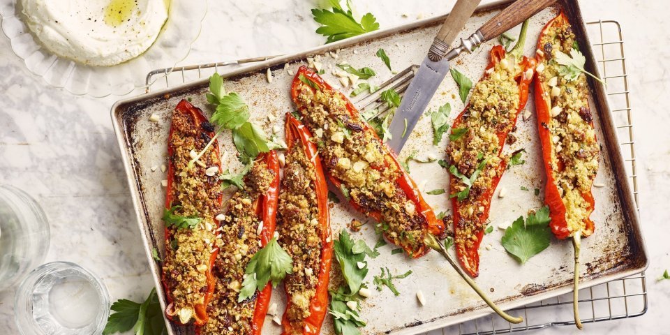 Gevulde puntpaprika's liggen op een metalen ovenschaal te schitteren. Ze zijn middendoorgesneden en opgevuld met couscous, gehakt en smaakmakers. Er liggen enkele blaadjes koriander bij voor extra kleur en het potje met homemade labneh is gepimpt met olijfolie en een draai van de pepermolen.