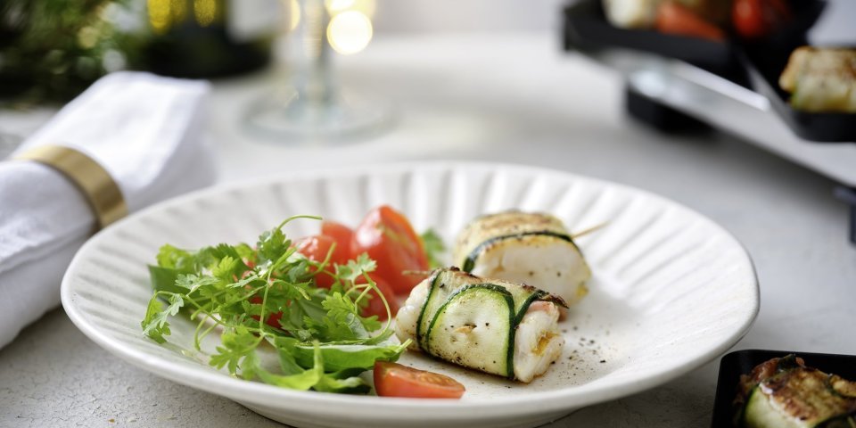 Twee pakjes liggen op een wit bord. Het pakje is gemaakt van een stukje vis verpakt in een sliertje courgette. We serveren dit feestelijk gerechtje met een frisgroene kruidensla en kertomaatjes.  