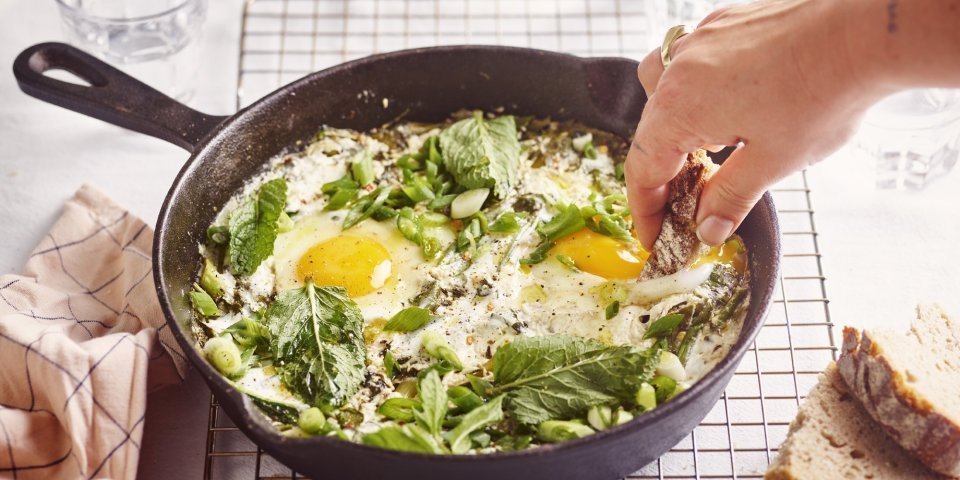 Op een rooster staat een zwarte gietijzeren pan met grieks getint gerechtje. Groene shakshuka met gebakken yoghurt. Er is een hand met een stukje brood in beeld dat er een dipje van zal nemen. 