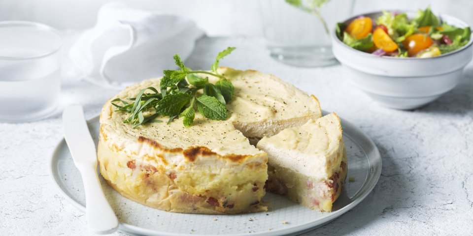 Een hartige cheesecake smaakt heerlijk als lunch of avondmaal. Door het tomatenslaatje krijg je de frisse toets die de cheesesmaak verfrissen. De cheescake ligt aangesneden op een groot rond bord, het slaatje staat langs het bord en een karaf water met takje munt zie je achteraan in de foto. 