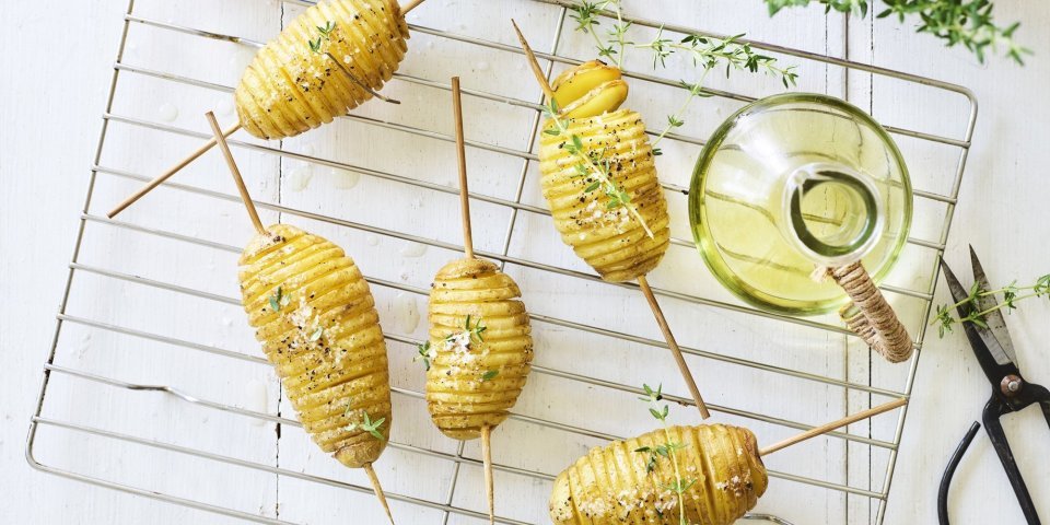 Hasselback potatoes liggen op een bakrooster te blinken. Ze zijn doorprikt met lange houten prikker om makkelijker te kunnen draaien op de BBQ. Er staat een flesje olie, verse tijm en een potje zeezout naast. 