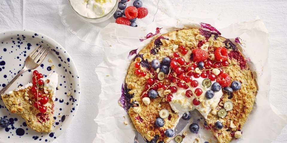 Een aangesneden taart staat op tafel met een wit laken er op. De kleuren van het rood fruit geven hierdoor nog een mooier zomers kleur. Naast de havermouttaart staat een bordje met blauwe spikkels en hier het stukje taart erop en een glaasje met de topping van de platte kaas en citroenzeste. 