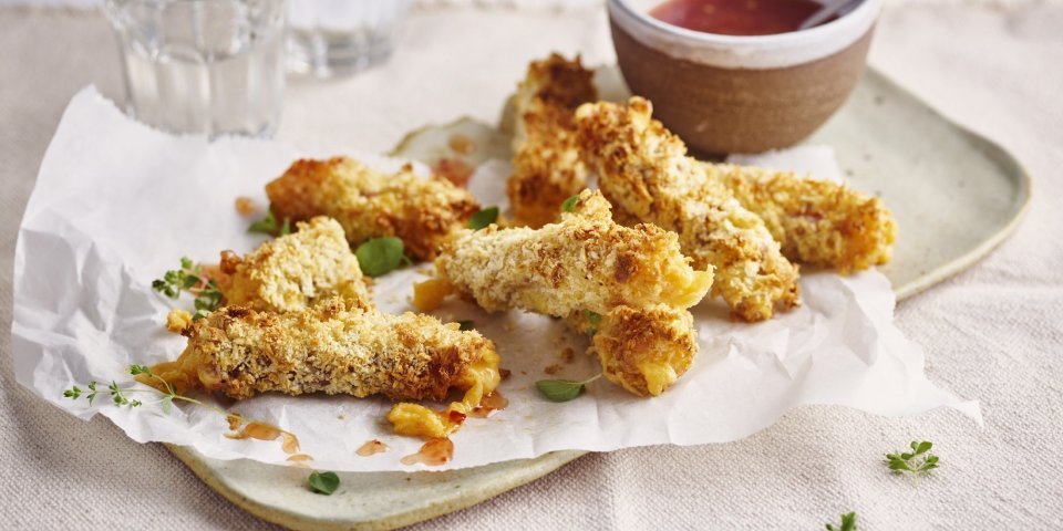 Krokante kaaskroketten liggen op een dienchaaltje, met een dipsausje in een zelfgedraaid keremakiekpotje. Er staan twee glaasjes water naast en de sticks zijn afgewerkt met verse oregano.