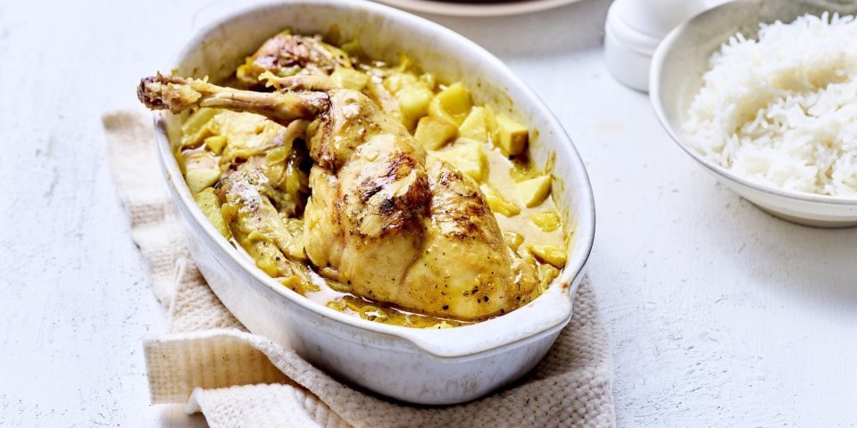 Op tafel staat een witte ovale ovenschotel met hierin de gele currysaus met stukjes appel en ananas. Hier bovenop liggen de bouten te blinken. De ovenschotel staat op een beige linnen handdoek met rijst, borden en een witte pepermolen langs.