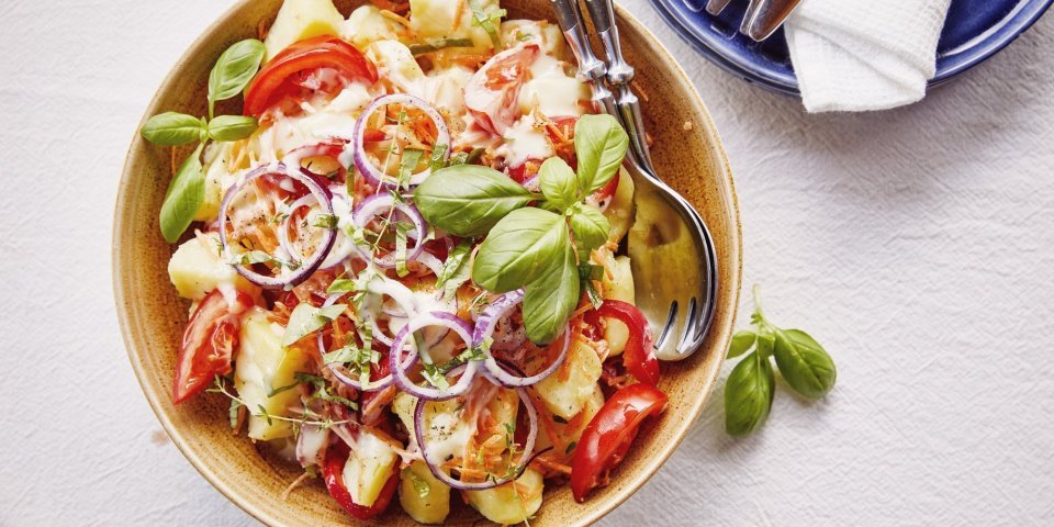 Een grote serveerschaal staat op de tafel, ze is gevuld met gekookte aardappelstukjes, tomaat, paparika, rode ui en wortel. De dressing is al toegevoegd en verse kruiden zoals basilicum en tijm geven een nog extra kleurtje aan de salade.