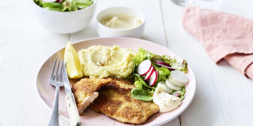 Een homemade shnitzel ligt op een pastelroze bord, samen met aardappelpuree, verse sla en zelfgemaakte mayonaise. Er is nog wat extra salade in een wit potje en mayonaise voor extra lekkers op te scheppen. 