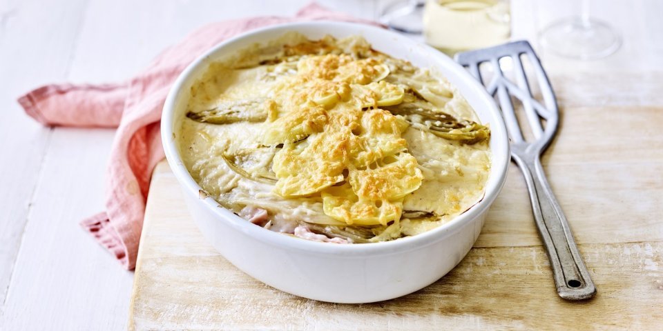 Er staat een ovenschotel op een houten plank, op de tafel. Er ligt een lepel naar de ovenschotel die gevuld is met een lasagne van witloof, ham, kaas en aardappelen. Er ligt ook een coraalroze servette bij.