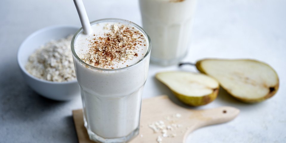 Twee klaargemaakte milkshakes staan op het lichtgrijze aanrecht te pronken. Er ligt een peer langs die doormidden gesneden is en wat havermoutvlokken gemorst. De shakes zijn afgewerkt met wat kaneelpoeder. 