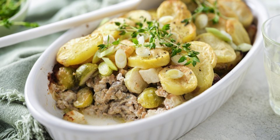 Een ovenschotel staat op het midden van de tafel, er is al een portie uitgeschept. Je ziet het gehakt onderaan liggen, met spruitjes , stukjes knolselder en goudgele aardappelschijfjes. 