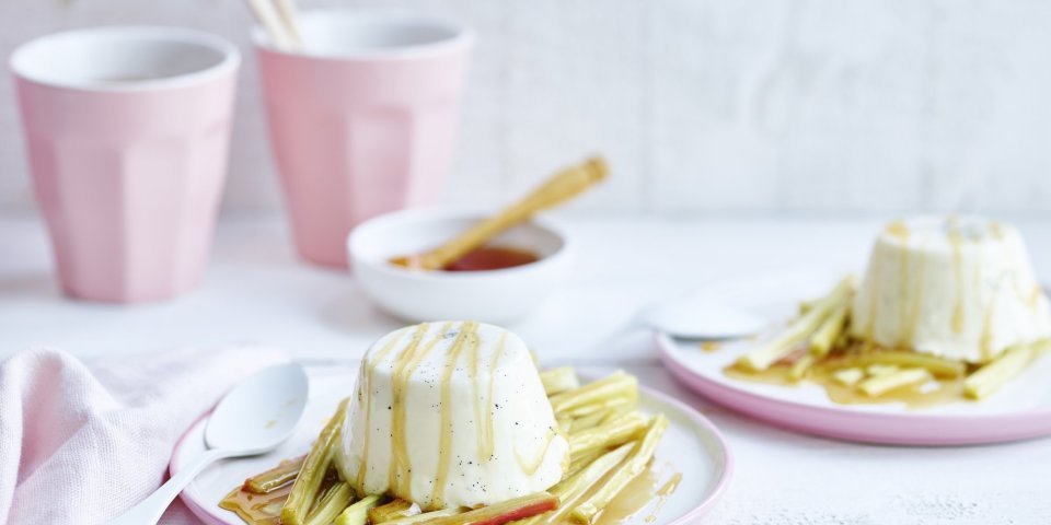 Een streling voor het oog, deze panna cotta met rabarber. Hij is opgediend in pastelroze bordjes en staat op een witte houten tafel. 