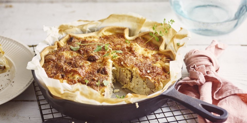 Op het aanrecht ligt een zwarte rooster met hierop de quiche in een gietijzeren pan. Hierbij ligt een roze servet, mes en bordje waarop al een stukje quiche op ligt. De quiche is afgewerkt met enkele verse blaadjes oregano. 