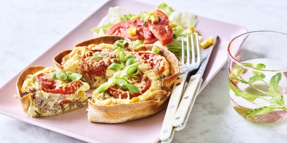 Een klaargemaakte quiche ligt op een roze en rechthoekig bord klaar om te eten. De quichebodem bestaat uit een wrap met hierin het quichebeslag en de groenten. 