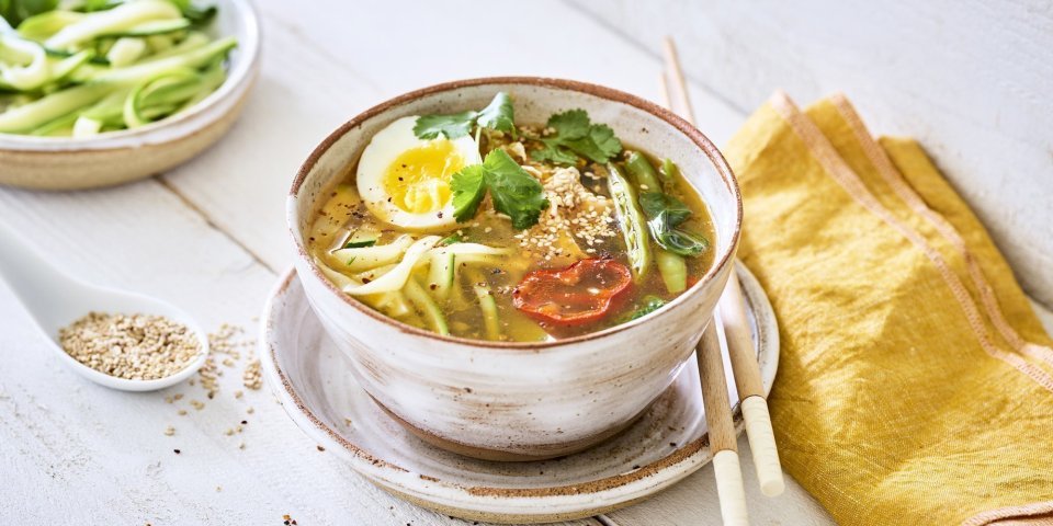 Een goeie portie soep staat uitgeschept op een houten witte tafel. De Japanse soep bestaat uit noedels van courgette aangevuld met peultjes, puntpaprika en kip. De soep is afgewerkt met een rood schijfje van een chilipeper en een gekookt eitje. 