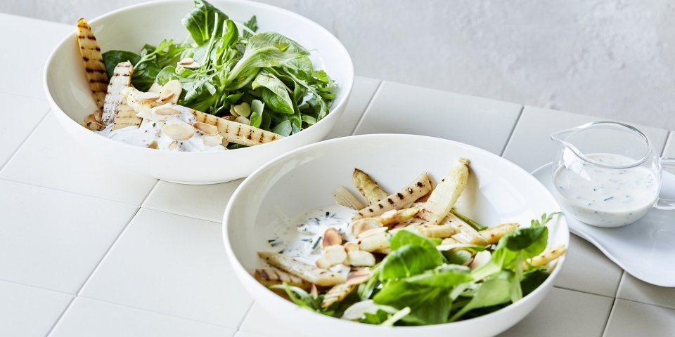 Salade met gegrilde asperges in 2 diepe borden. De groene salade bestaat uit veldsla en rucola met daarbovenop gegrilde witte asperges. Er is ook een yoghurtdressing met verse kruiden die bovenaan het bord staat in een glazen karafje. 