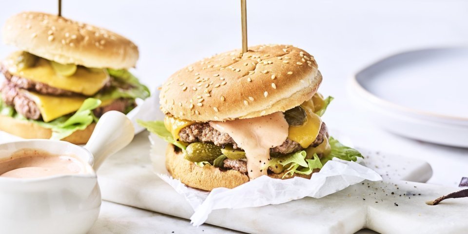 Twee heerlijke burgers liggen klaar op een marmere snijplank. Het vlees is verdeeld in twee dunne porties met de saus en gesmolten kaas er bovenop. De burgers worden recht gehouden door middel van een prikker.