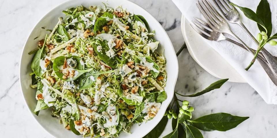 Een gezonde side dish voor je feestgerecht dat extra stukje power te geven. Deze spruitjessalade is opgediend in een witte keramische kom met stukjes appel en afgewerkt met een dressing op basis van blauwekaas, zure room en mayonaise. Er ligt bestek langs op een witte nap. 