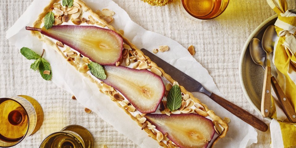 Op tafel ligt een heerlijke rechthoekige perentaart. De bodem is gevuld met verse kwark en hier bovenop liggen de gestoofde peren. De taart is afgewerkt met gegrilde amandelschilfers. 