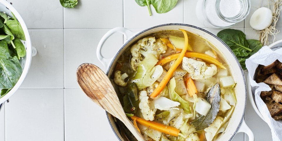 Een lentestoofpotje boordevol groenten, denk maar aan bloemkool, wortel, koolrabi, prei en spinazie. De stoofpot staat op het aanrecht in een grote kookpot, rond de pot liggen nog enkele spinazieblaadjes en een flesje room.