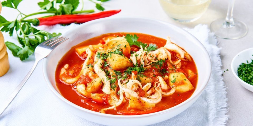 Een tomatenrood stoofpotje met vis staat op tafel. Het is een stoofpotje met stukjes paprika, aardappelen en tomatensaus. 