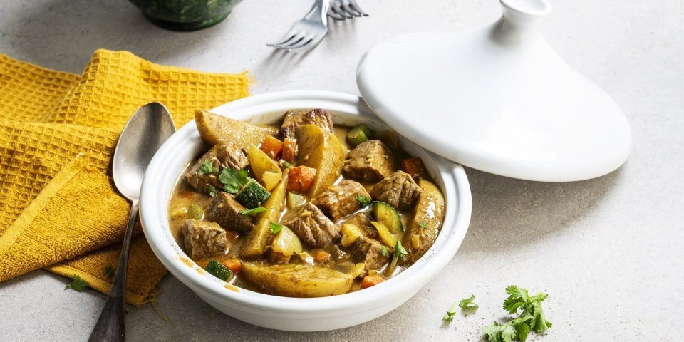 Op tafel staat een witte tajine met een gerechtje van rundvlees, courgette, selder, wortel en stukjes aardappel. Er ligt een okerkleurige servette bij en groene kommetjes om de tajine in op te scheppen. 