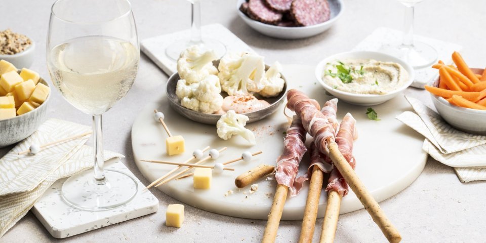 Een mooie marmere serveerplank in rond vorm staat op tafel. Hij is gevuld met allerlei tappas, zoals bloemkoolroosjes met cocktailsaus, wortel, kaasblokjes, salami met mosterd en homemade hummus.