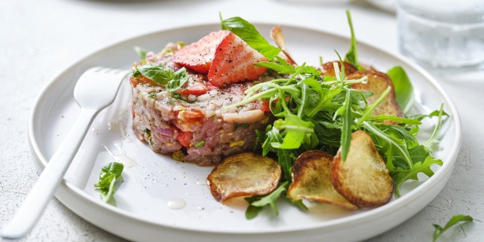 Zoek je een alternatief voor steak-tartaar? Wel dit is een zomerversie, nl eentje met kalfsvlees, aardbeien, tomaatjes en verse kruiden. We werkten het gerechtje af met gebakken aardappelplakjes en verse rucola. Er ligt een wit vork en servette naast om meteen aan te vallen. 