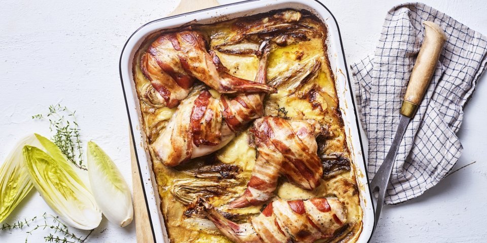 Op een grote houten snijplank staat de ovenschaal met de tartiflette en stukken konijn. Het konijn in omwikkeld met sneetjes spek, waardoor het nog iets extra heeft. Er ligt witloof langs en verse tijm.