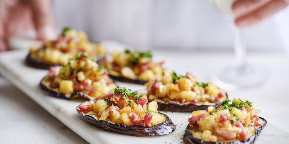 Op een marmere serveerschaal liggen enkele aubergineplakjes. Deze dienen als toastje en zijn belegd met stukjes Berloumi en spek. 