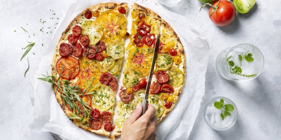 Een kleurrijke tomatentaart staat op tafel op een wit boterpapiertje. Ze wordt aangesneden door een onbekende vrouw met een koksmes. Rechtsboven liggen nog enkele tomaten om ze extra in de picture te zetten. 