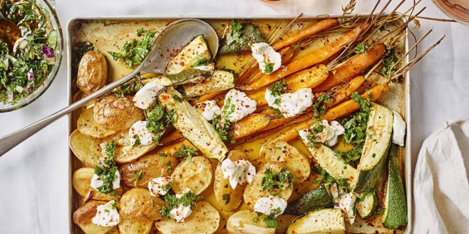 Ovenplaat met gegrilde groentjes komt net uit de oven en staat al op tafel. Hij heeft warme kleuren van de gouden aardappelen naar de oranje wortelen. Hij is afgewerkt met toefjes geitenkaas en verse chimichurri.